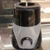 An image of a compact electric mixer grinder or blender base resting on a polished glass-top counter in an indoor showroom or workshop environment. The appliance features a dual-tone plastic body, with a glossy black upper and lower section split by a swooping, V-shaped light grey accent panel across the front face.