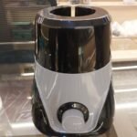 An image of a compact electric mixer grinder or blender base resting on a polished glass-top counter in an indoor showroom or workshop environment. The appliance features a dual-tone plastic body, with a glossy black upper and lower section split by a swooping, V-shaped light grey accent panel across the front face.