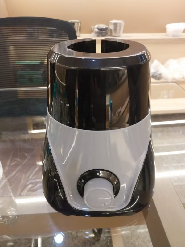 An image of a compact electric mixer grinder or blender base resting on a polished glass-top counter in an indoor showroom or workshop environment. The appliance features a dual-tone plastic body, with a glossy black upper and lower section split by a swooping, V-shaped light grey accent panel across the front face.