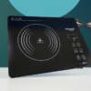 A high-definition, sharp studio product photograph of a sleek, black portable induction cooktop sitting on a clean white surface. The cooktop features a minimalist design with a large circular heating zone marked by concentric white rings and a central warning symbol triangle.