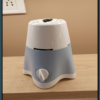 A crisp, close-up photograph of a compact electric mixer grinder base resting on a light-colored wooden countertop against a neutral beige wall. The appliance features a two-tone plastic body with a glossy white upper section and a matte, light-blue lower section that tapers down into a stable four-legged clover base with a white bottom trim.